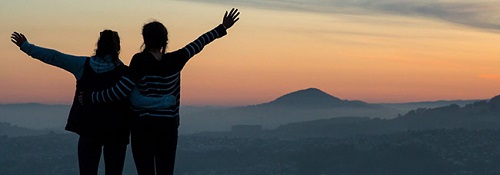 Two girls on top of a mountain at sunset, arms around each other and raised into the air. 